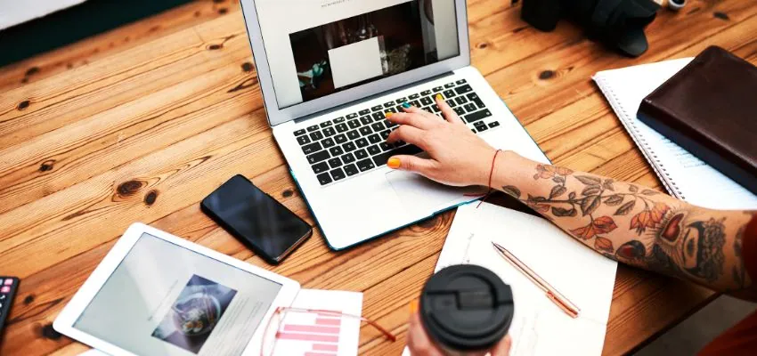 A person working on a laptop at a wooden desk, surrounded by a tablet, smartphone, notebook, and coffee cup, illustrating the creative process behind writing blogs with Jasper AI.